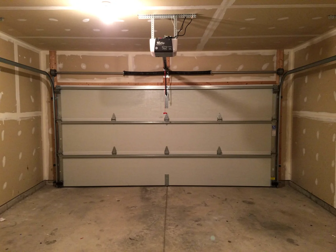 A white garage door is closed inside a garage with unfinished drywall.